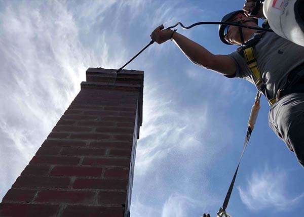 Masonry repairs-Tech cleaning tall chimney wearing safety gear with wispy clouds in the background - C&C Chimney